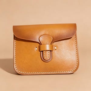 May include: A small, rectangular, tan leather bag with a flap closure and a buckle. The bag has a stitched border and a lighter brown hue on the flap. The bag is designed with a simple, classic aesthetic.