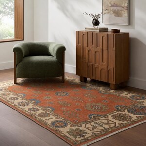 May include: A room setting featuring a rust-colored area rug with a beige border and intricate geometric patterns. A green armchair with wooden legs sits near a wooden cabinet. The room has a neutral color palette.