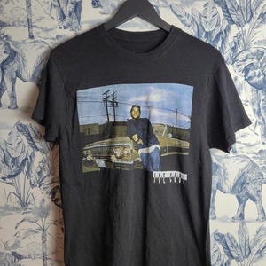 Pode incluir: Camiseta preta com um gráfico de uma pessoa encostada em um carro clássico. A imagem tem um fundo de céu azul e o texto "Ice Cube" em branco. A camiseta está pendurada em um cabide preto.