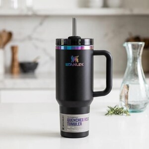 Op de afbeelding: Een zwarte Stanley Quencher H2.0 tumbler met een handvat en een rietje. De tumbler heeft een regenboogkleurige rand en het Stanley-logo. Het label luidt "Quencher H2.0 Tumbler" en "Black Chrome Collection."