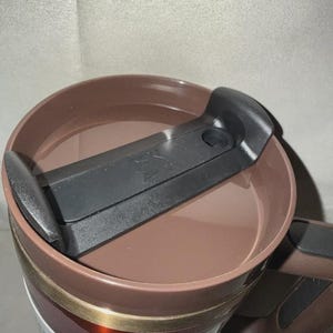 May include: A brown travel mug with a black lid and handle. The lid has a sliding mechanism to open and close the drinking spout. The mug has a white label with the text "H2.0".