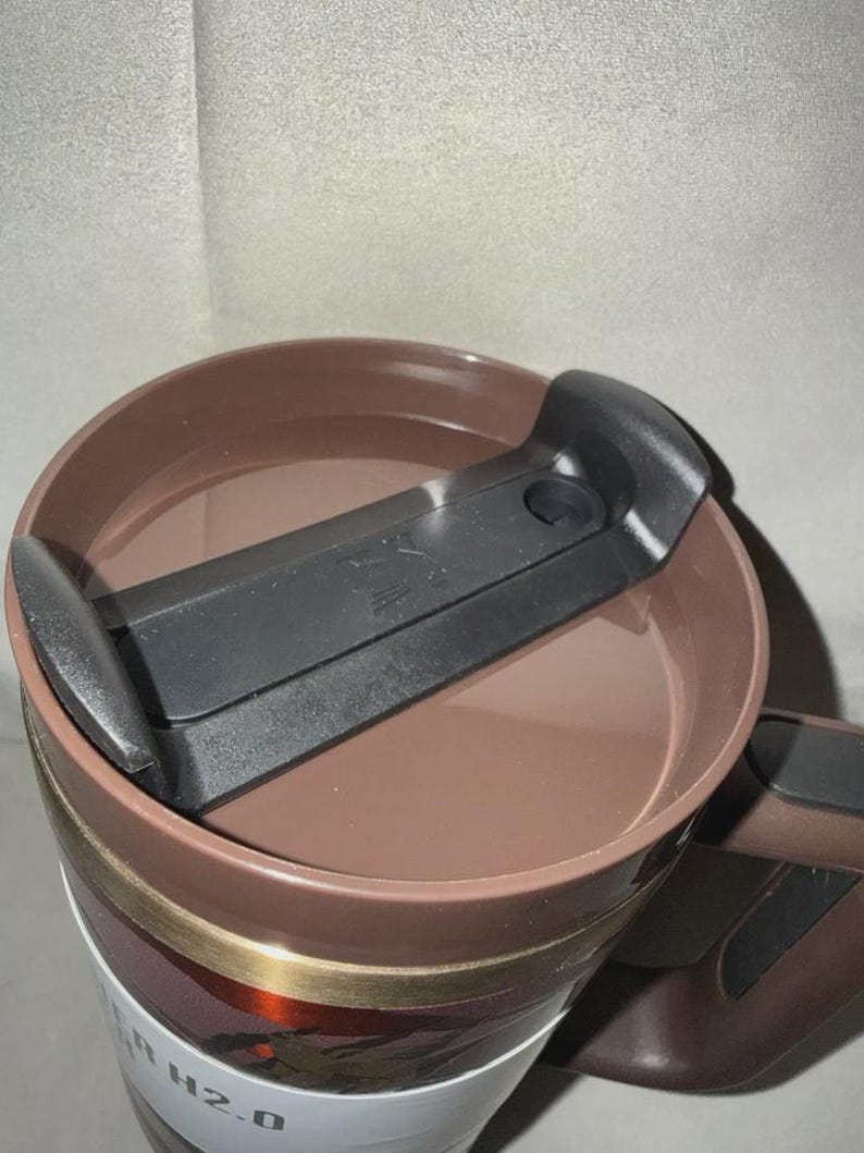 May include: A brown travel mug with a black lid and handle. The lid has a sliding mechanism to open and close the drinking spout. The mug has a white label with the text "H2.0".