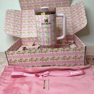 May include: A Stanley LoveShackFancy tumbler with a pink and green floral pattern, a clear lid, and a light pink handle, presented in a matching floral box with a pink fabric pouch featuring a pink ribbon printed with Stanley LoveShackFancy text.