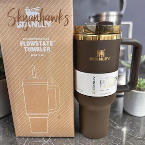 May include: A brown and gold Stanley Flowstate tumbler with a handle and a straw, next to its matching box. The box is brown with the text "Skyanhawks" and "The Quencher H2.0 Flowstate Tumbler 40 oz | 1.18 L".