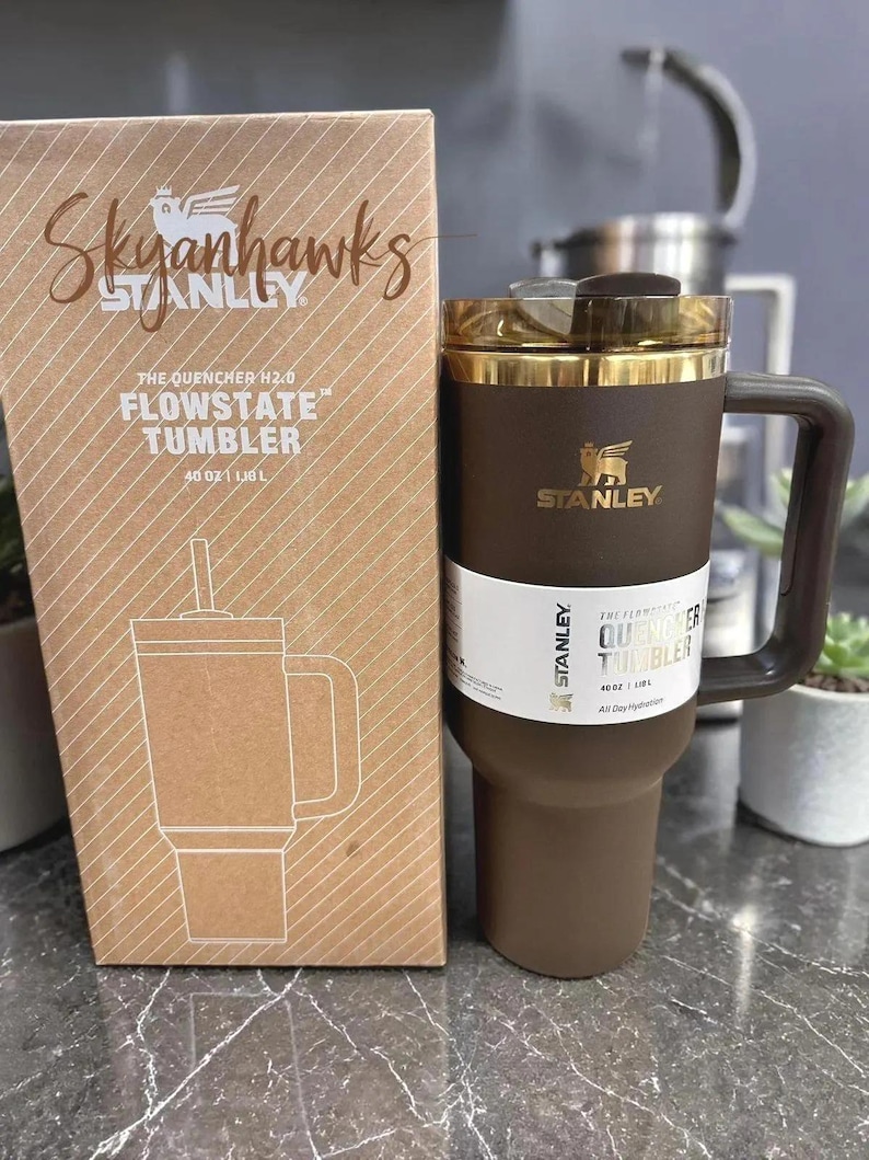 May include: A brown and gold Stanley Flowstate tumbler with a handle and a straw, next to its matching box. The box is brown with the text "Skyanhawks" and "The Quencher H2.0 Flowstate Tumbler 40 oz | 1.18 L".