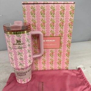 May include: A pink Stanley Quencher H2.0 tumbler featuring a floral pattern, a matching box with the text Stanley LoveShackFancy, and a pink drawstring pouch. The tumbler has a clear lid, a gold-tone rim, and a side handle with floral print.