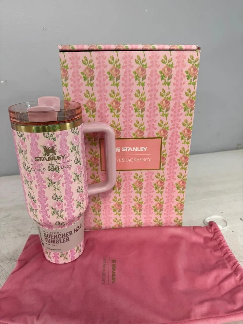 May include: A pink Stanley Quencher H2.0 tumbler featuring a floral pattern, a matching box with the text Stanley LoveShackFancy, and a pink drawstring pouch. The tumbler has a clear lid, a gold-tone rim, and a side handle with floral print.
