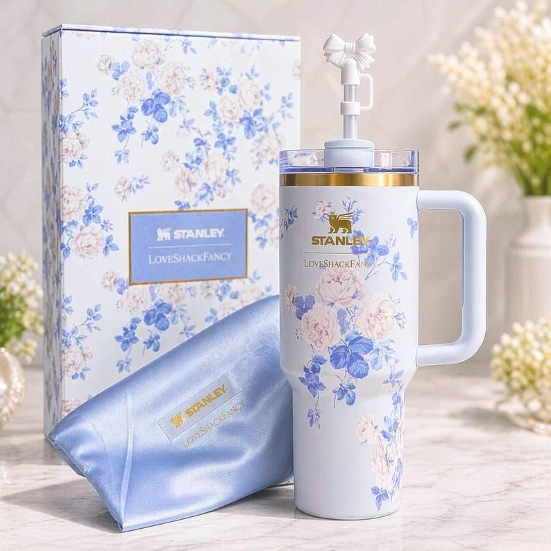 Puede incluir: Un vaso Stanley azul claro con asa, adornado con un estampado floral de rosas rosas y azules. El vaso tiene un lazo blanco en la pajita y detalles dorados. Tambi&eacute;n hay una caja de regalo y una bolsa de sat&eacute;n a juego, ambas con el mismo dise&ntilde;o floral y el texto "Stanley LovesShackFancy".