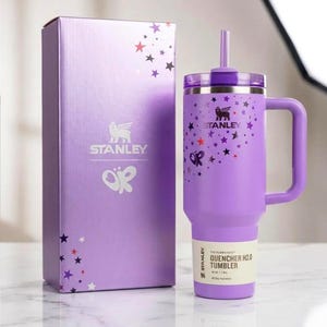 以下が含まれることがあります： ハンドル、ストロー、蓋が付いた紫色のスタンレーQuencher H2.0タンブラーと、お揃いの箱。タンブラーと箱は星と蝶のグラフィックで装飾されています。タンブラーには「THE FLOWSTATE QUENCHER H2.0 TUMBLER」と「40 OZ / 1.18L」の文字があります。