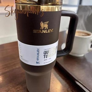 May include: A dark brown Stanley Quencher Tumbler with a gold-colored rim and lid. The tumbler has a black handle and a white label with text. The Stanley logo is visible. The tumbler holds 40 oz.