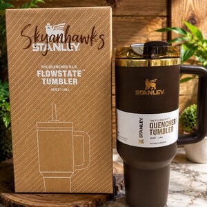 May include: A brown 40 oz Stanley Flowstate Quencher tumbler with a handle and a clear lid, accompanied by its matching cardboard packaging box. The box features the text Skyanhawks, The Quencher H2.0 Flowstate Tumbler, and 40 oz | 1.18 L.