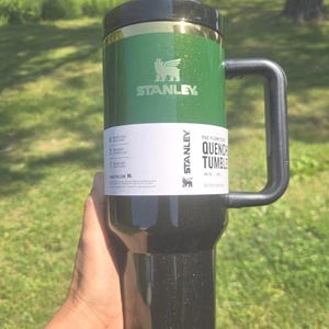 May include: Green and black Stanley Quencher Tumbler with a handle. The tumbler has a black lid and a gold band. The Stanley logo is visible on the green section. The tumbler is 40 oz.