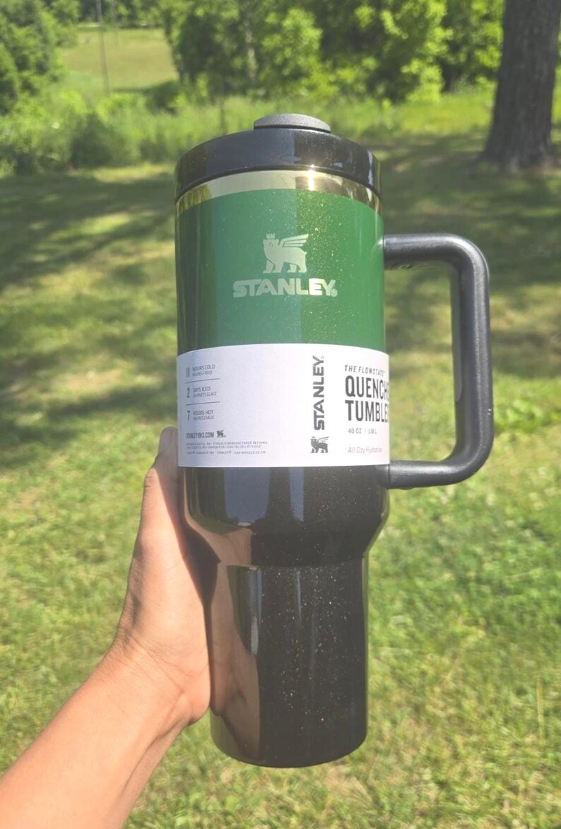 May include: Green and black Stanley Quencher Tumbler with a handle. The tumbler has a black lid and a gold band. The Stanley logo is visible on the green section. The tumbler is 40 oz.