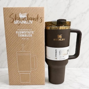 May include: A dark brown Stanley Quencher H2.0 Flowstate tumbler with a 40 oz capacity and a matching handle, positioned next to its cardboard packaging box which features the text Skyhawks, Stanley, and The Quencher H2.0 Flowstate Tumbler 40 oz 1.18 L.