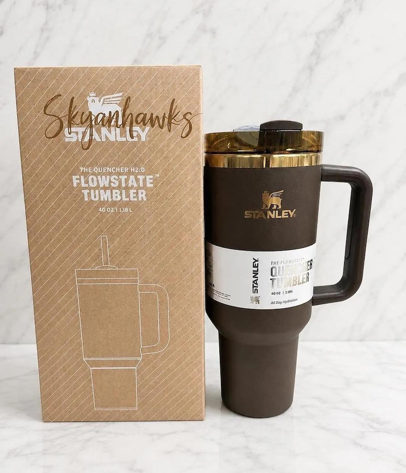 May include: A dark brown Stanley Quencher H2.0 Flowstate tumbler with a 40 oz capacity and a matching handle, positioned next to its cardboard packaging box which features the text Skyhawks, Stanley, and The Quencher H2.0 Flowstate Tumbler 40 oz 1.18 L.