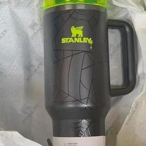 May include: A black Stanley tumbler with a neon green lid and matching logo. The body features a textured spiderweb pattern. A white product label at the base displays text including 11 HOURS COLD and 2 DAYS ICED. It has a black side handle.