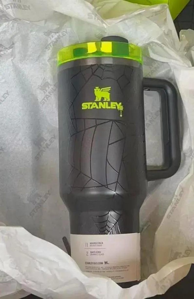 May include: A black Stanley tumbler with a neon green lid and matching logo. The body features a textured spiderweb pattern. A white product label at the base displays text including 11 HOURS COLD and 2 DAYS ICED. It has a black side handle.