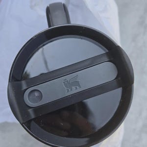 May include: A black travel mug with a handle and a lid. The lid has a sliding closure and a small, embossed logo of a flying creature. The mug is designed for hot or cold beverages.