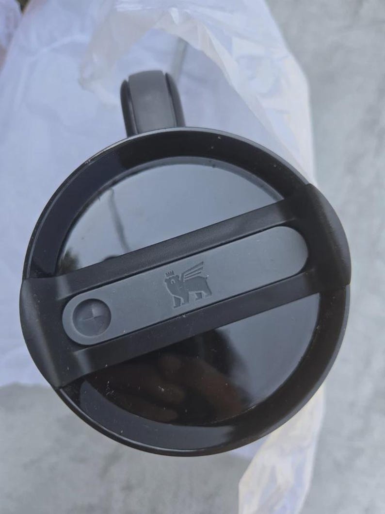 May include: A black travel mug with a handle and a lid. The lid has a sliding closure and a small, embossed logo of a flying creature. The mug is designed for hot or cold beverages.
