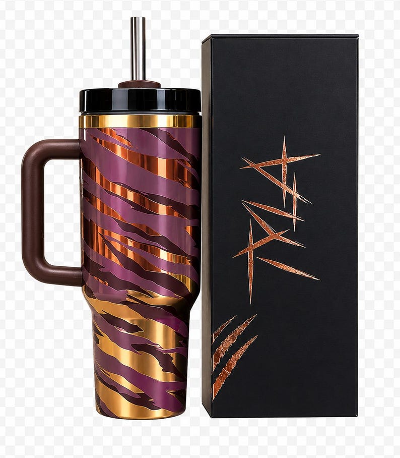 May include: A stainless steel tumbler with a handle, straw, and lid. The tumbler has a purple and gold tiger stripe pattern. A black box with copper-colored text and claw marks is next to the tumbler.