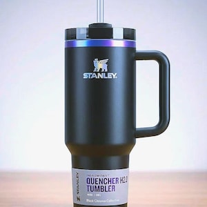 Op de afbeelding: Zwarte Stanley Quencher H2.0 Tumbler met een handvat en een rietje. De tumbler heeft een zwarte behuizing met een zilveren en iriserende rand. Het Stanley-logo is zichtbaar op de voorkant. De tekst "QUENCHER H2.0 TUMBLER" staat onderaan.