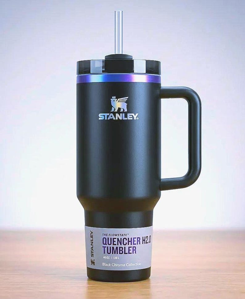 Op de afbeelding: Zwarte Stanley Quencher H2.0 Tumbler met een handvat en een rietje. De tumbler heeft een zwarte behuizing met een zilveren en iriserende rand. Het Stanley-logo is zichtbaar op de voorkant. De tekst "QUENCHER H2.0 TUMBLER" staat onderaan.