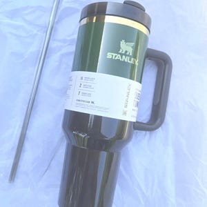 May include: Green and black Stanley travel mug with a handle and a silver straw. The mug has the Stanley logo and a label with text. The mug is designed to keep drinks cold for 11 hours and hot for 7 hours.