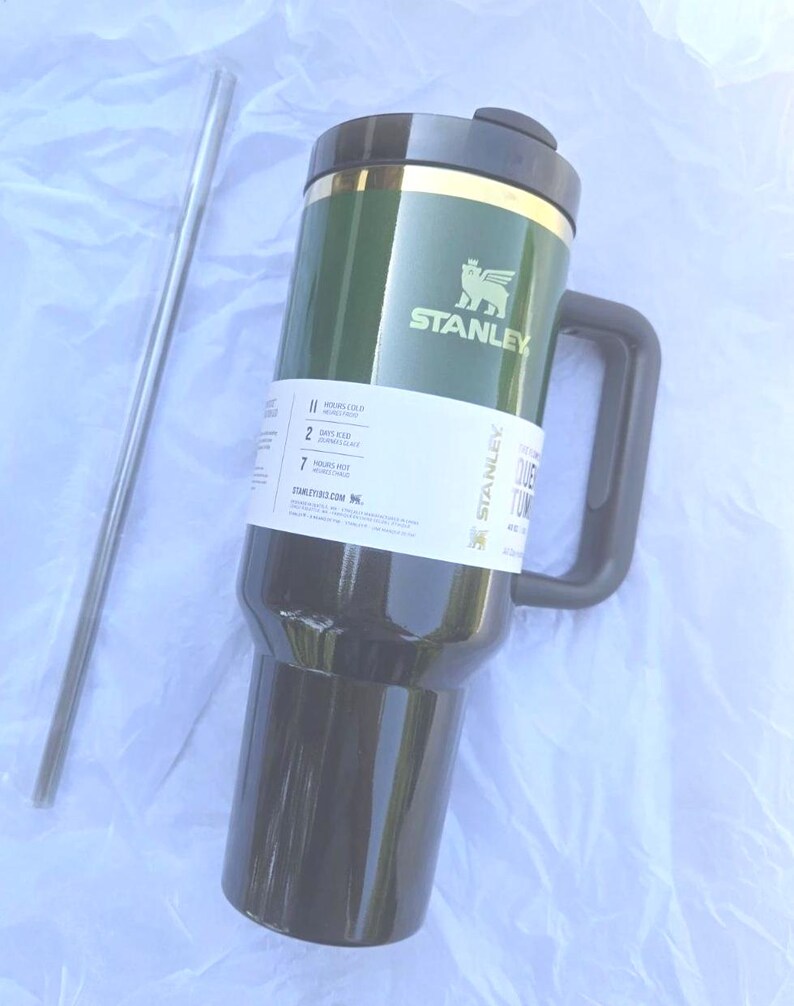 May include: Green and black Stanley travel mug with a handle and a silver straw. The mug has the Stanley logo and a label with text. The mug is designed to keep drinks cold for 11 hours and hot for 7 hours.