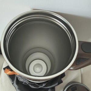 May include: A stainless steel insulated travel mug with a brown handle and lid. The interior is silver, and the mug sits on a black satin bag. An orange ribbon is tied around the top.