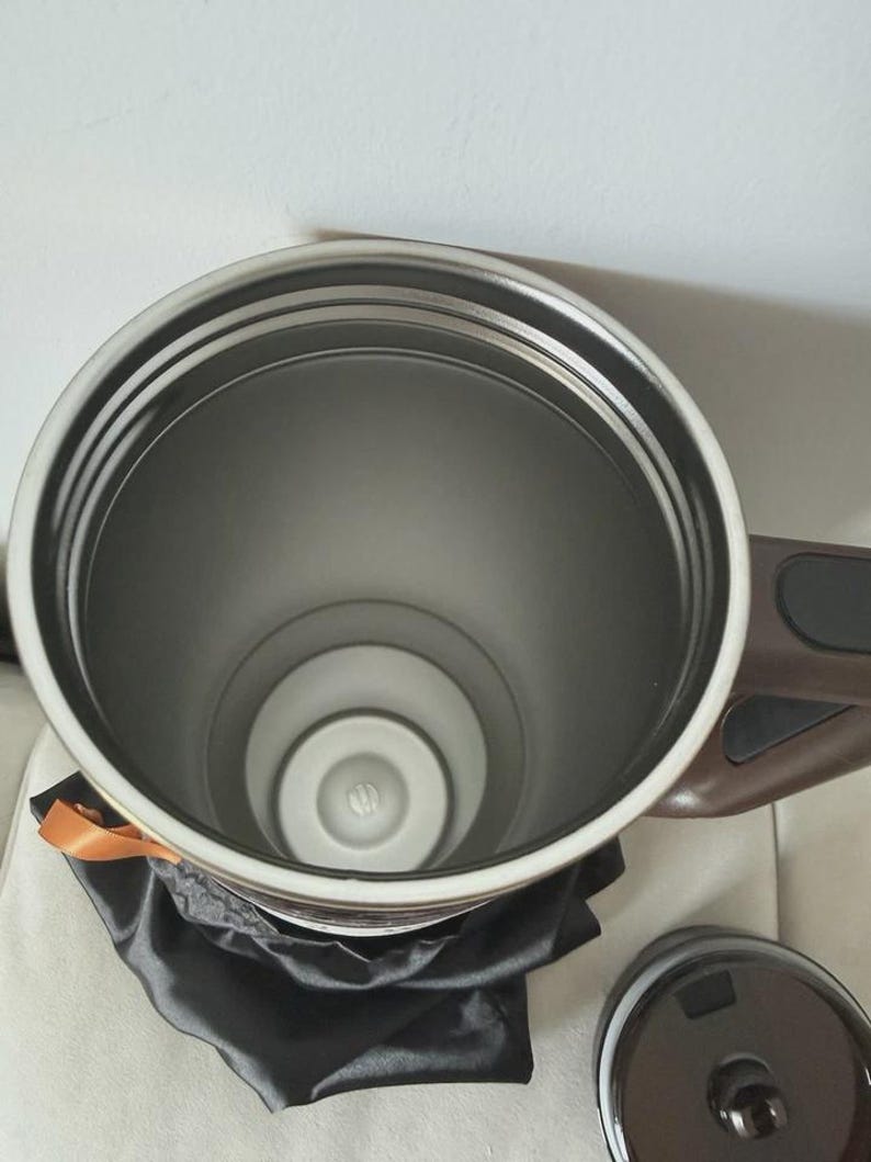 May include: A stainless steel insulated travel mug with a brown handle and lid. The interior is silver, and the mug sits on a black satin bag. An orange ribbon is tied around the top.