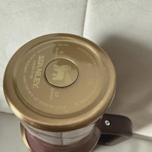 May include: A Stanley brand travel mug with a brown handle and a gold-colored lid. The lid has the Stanley logo and text that reads "A Brand of PMI" and "Do Not Microwave". The mug is a mix of brown and mauve.