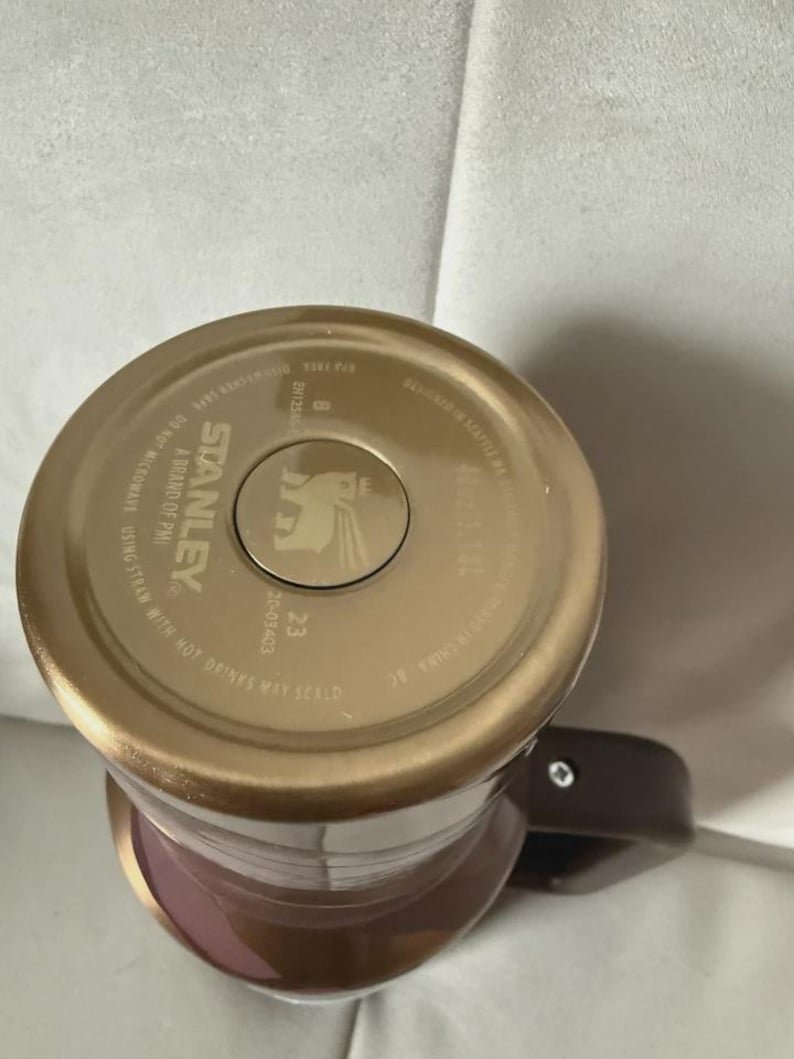 May include: A Stanley brand travel mug with a brown handle and a gold-colored lid. The lid has the Stanley logo and text that reads "A Brand of PMI" and "Do Not Microwave". The mug is a mix of brown and mauve.