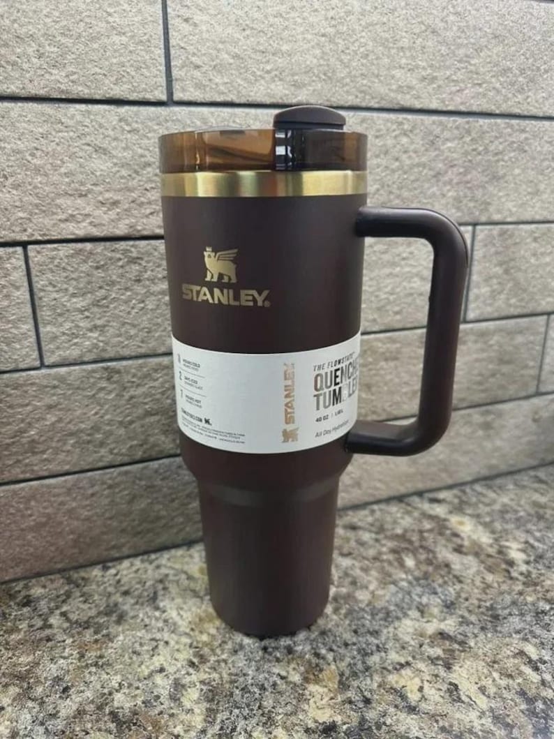 May include: A dark brown Stanley Quencher tumbler with a handle and a clear lid. The tumbler has a gold band around the top and the Stanley logo in gold. A white label with text is wrapped around the tumbler.