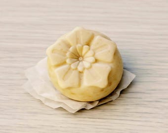 May include: A round, off-white, textured soap bar featuring a molded floral design on its top surface. The soap rests on a small, crinkled piece of white paper, showcasing a smooth, slightly speckled appearance with a detailed, symmetrical flower pattern.