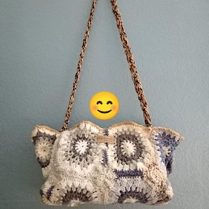 Può includere: Una borsa all'uncinetto fatta a mano con un design patchwork nei toni del bianco, grigio e blu. La borsa ha una tracolla marrone e beige e una piccola etichetta con la scritta "Handmade". La borsa è appesa a una parete azzurra.