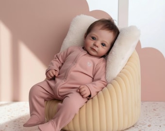 May include: A realistic baby doll with brown hair and blue eyes, wearing a long-sleeved, footed, dusty pink cotton onesie with a small white embroidered heart on the chest, seated in a cream-colored, ribbed, curved, upholstered chair.