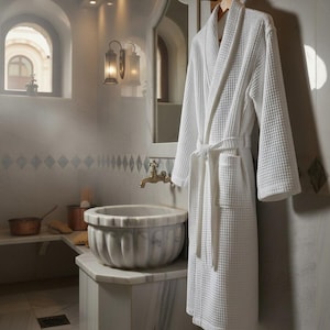 May include: A white waffle-weave bathrobe hangs on a wooden rack in a luxurious bathroom. The robe has a shawl collar, a tie belt, and pockets. The bathroom features a marble sink, a brass faucet, and a decorative wall sconce.
