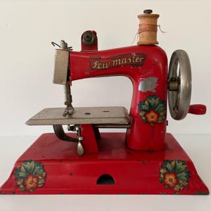 May include: A vintage red Sew Master sewing machine featuring a silver metal hand crank, a needle bar, and a flat work surface. The body is decorated with floral decals on the base and side, and a wooden spool sits on the top spindle.