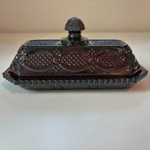 May include: A rectangular dark red glass butter dish with a matching lid. The lid features a textured diamond pattern, oval borders, and a central knob shaped like an acorn. The base has a scalloped edge with a repeating geometric pattern.