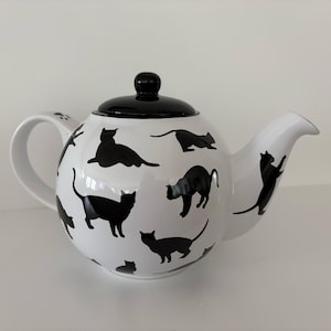 May include: A white ceramic teapot featuring a pattern of black cat silhouettes in various poses. The teapot has a rounded body, a curved handle with a small paw print detail, a spout, and a removable solid black lid with a round knob handle.