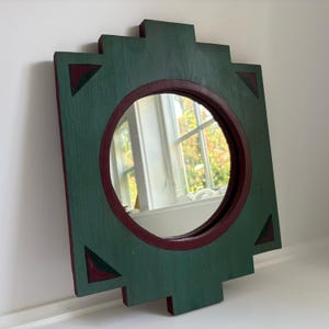 May include: A square wooden mirror with a stepped, geometric frame painted in dark green. The circular mirror glass is set within a deep maroon border. Four triangular cutouts in the corners reveal the same maroon color underneath the green.