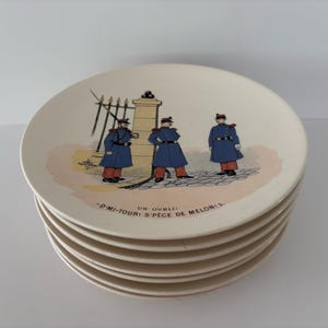 May include: A stack of cream-colored ceramic plates featuring a printed illustration of three soldiers in blue uniforms with red accents standing near a stone gate. The text "UN OUBLI! -D'MI-TOUR! S'PÈCE DE MELON!S." is printed below the soldiers.