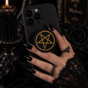 May include: A black smartphone grip featuring a gold pentagram design centered on a circular black background. The grip is attached to the back of a black smartphone, showcasing a clean, circular aesthetic with a bold, contrasting gold star symbol.