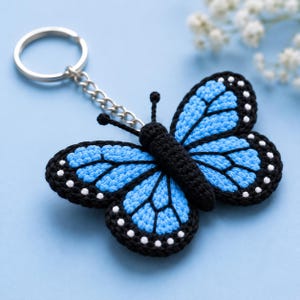 May include: A crocheted butterfly keyring with a silver ring. The butterfly has light blue wings with black outlines and white dots. The body is black. The keyring is set against a light blue backdrop.
