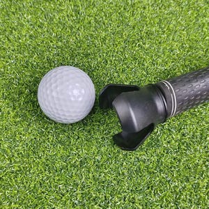 Könnte beinhalten: Ein weißer Golfball und ein schwarzer Golfschlägergriff mit einem Ballfangaufsatz liegen auf einer grünen Kunstrasenoberfläche. Der Golfball hat eine strukturierte Oberfläche.