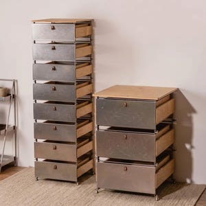 May include: Two metal drawer chests with wooden tops and pulls. One chest has ten drawers, and the other has three. The drawers are partially open, revealing the wooden interiors. The chests sit on a neutral-colored rug.