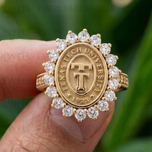 May include: A gold ring featuring an oval-shaped center plate with the embossed Texas Tech University logo and the year 1923. The central emblem is surrounded by a halo of small, round, clear white stones set in a scalloped gold frame.