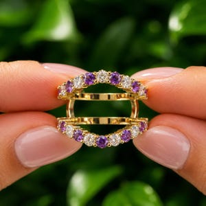 May include: A gold ring guard featuring two parallel bands connected by vertical bars. The bands are set with alternating round-cut clear white stones and round-cut purple stones in a prong setting, creating a continuous sparkling design.