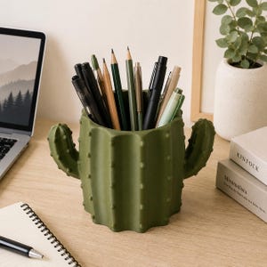 Peut inclure: Un porte-crayons vert olive mat en forme de cactus avec deux bras latéraux, rempli de divers stylos et crayons. Le récipient cylindrique présente une surface texturée avec de petites bosses en relief, servant d'organisateur de bureau fonctionnel.