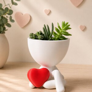 May include: A white ceramic planter shaped like a person, holding a red heart. The planter is filled with various green succulents. Decorative pink heart shapes adorn the background, creating a charming and romantic aesthetic.